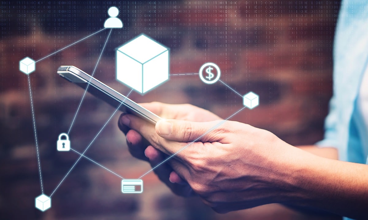 Imagem conceitual de tokenização, mostrando uma mão segurando um cubo digital brilhante no centro, representando tecnologia blockchain. Em torno do cubo, ícones relacionados a finanças, segurança e metas, como cadeado, cifrões e gráficos, conectados por linhas que simbolizam interconexão digital