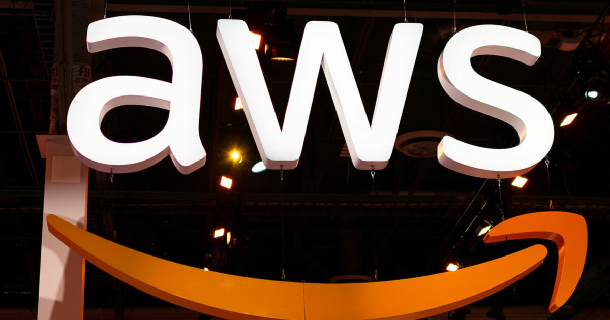 Imagem com o letreiro iluminado da sigla “AWS”, que representa a Amazon Web Services, pendurado no teto de um evento ou feira. Abaixo das letras está parcialmente visível a seta curvada laranja característica do logotipo da Amazon. Ao fundo, é possível ver refletores acesos e estruturas metálicas do ambiente interno (chile)