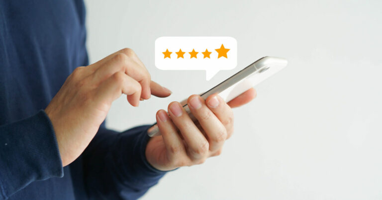 Imagem de uma pessoa segurando um smartphone com uma das mãos enquanto utiliza a outra para tocar na tela. Acima do dispositivo, há um balão de diálogo com cinco estrelas douradas, representando uma avaliação de alta qualidade ou feedback positivo. O fundo é branco, destacando a interação com o aparelho (clientes, experiência do cliente, CX)