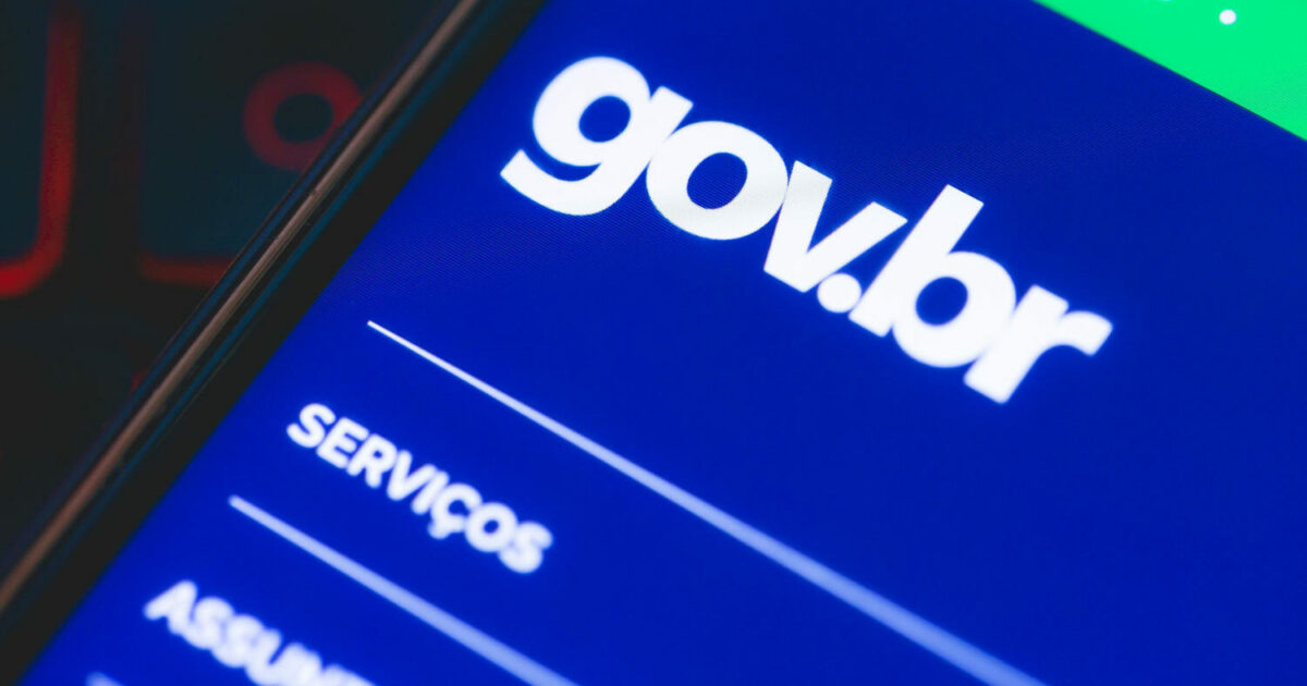 A imagem mostra a tela de um smartphone exibindo o aplicativo ou site do gov.br, a plataforma oficial de serviços digitais do governo brasileiro. A interface tem um fundo azul com texto branco, destacando o logo "gov.br" no topo e opções de navegação, como "SERVIÇOS", logo abaixo. O aparelho está inclinado, e há um fundo desfocado com tons vermelhos e pretos (gov.br, governo federal, tendências, contas)