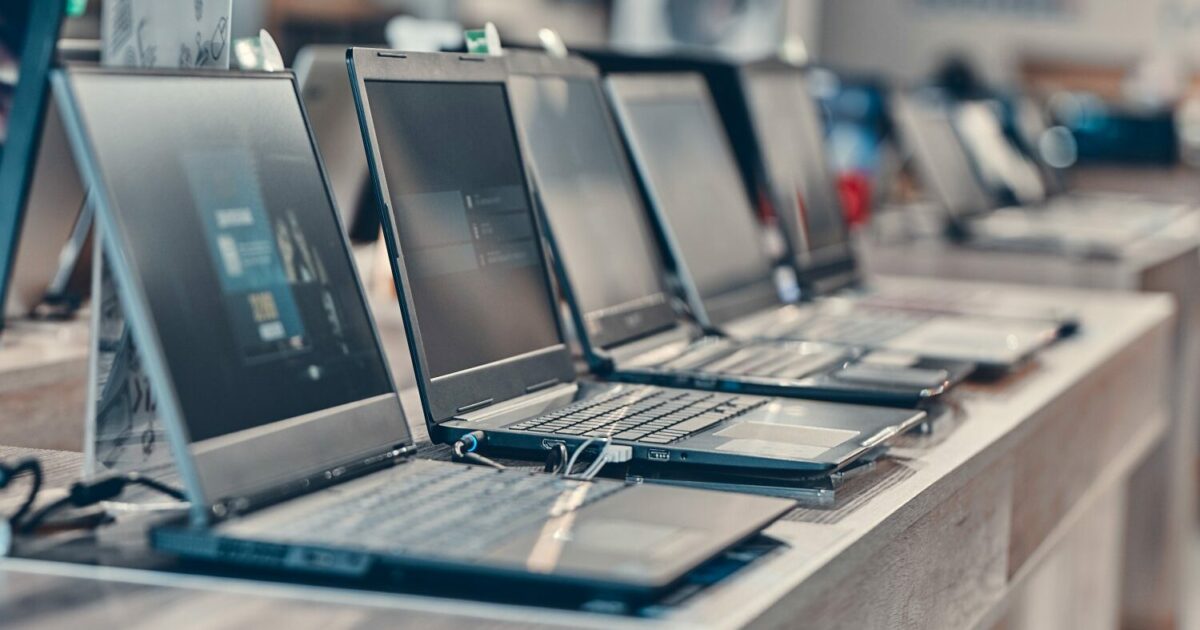 A imagem mostra uma fileira de notebooks expostos em uma loja de eletrônicos. Os laptops estão inclinados para frente em uma superfície de exposição, com as telas desligadas e os teclados visíveis. Cada dispositivo está conectado a cabos, possivelmente para fins de segurança ou demonstração. O ambiente é bem iluminado, e o foco está nos notebooks, com um fundo desfocado que indica um espaço de loja (notebooks, laptops, computadores, pcs, tarifas)