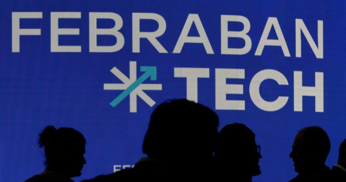 Imagem de um painel azul com o logotipo e o texto "FEBRABAN TECH" em destaque. Em frente ao painel, aparecem as silhuetas de várias pessoas, em clima de evento ou conferência, sugerindo um ambiente de tecnologia e negócios. (Febraban Tech Bancos 2025)