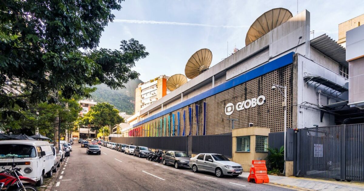 Fachada do prédio da emissora Globo, com logotipo visível na lateral do edifício. A construção é moderna, com detalhes em concreto e elementos verticais coloridos na parede lateral. Há várias antenas parabólicas no topo do prédio. A rua em frente está repleta de carros estacionados e algumas árvores proporcionam sombra. Ao fundo, é possível ver morros e vegetação típica do Rio de Janeiro. (globo, tv globo)