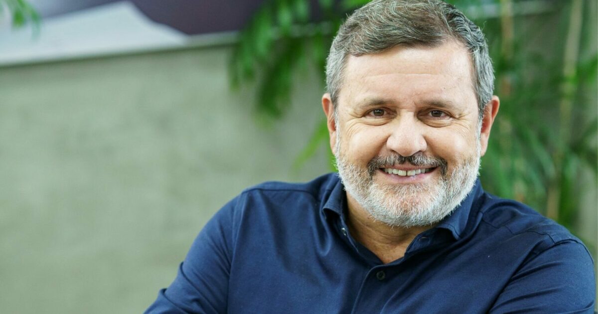 Vittorio Danesi, CEO da Simpress, sorrindo para a câmera. Ele é um homem de meia-idade com barba grisalha e cabelo curto, vestindo uma camisa azul escura. O fundo da imagem é levemente desfocado, com elementos verdes de plantas e uma parede neutra