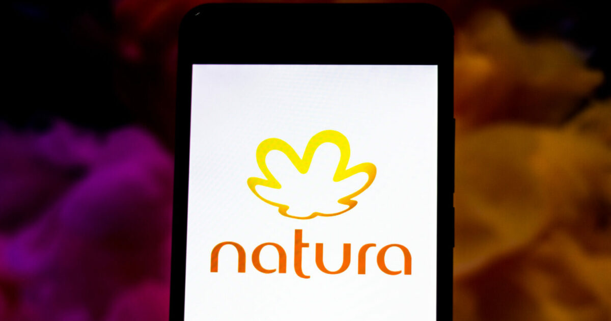 Tela de um smartphone exibindo o logotipo da empresa Natura, com um fundo branco e uma figura estilizada em forma de folha na cor amarela acima da palavra "natura" escrita em letras minúsculas laranja. Ao fundo, há um desfoque com tons escuros e coloridos, predominando rosa, roxo e amarelo. Natura (consultor)