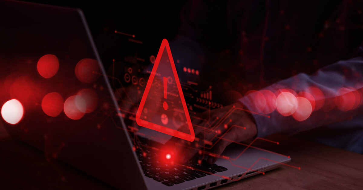 Imagem sombria com um laptop em foco e um símbolo de alerta vermelho no centro, representando uma ameaça digital. Há uma sobreposição de gráficos digitais abstratos em vermelho, sugerindo atividades cibernéticas ou ataques de malware. As mãos de uma pessoa digitando no teclado são visíveis ao fundo, enfatizando o risco de segurança online (malwares, ScarletStealer, Eset)