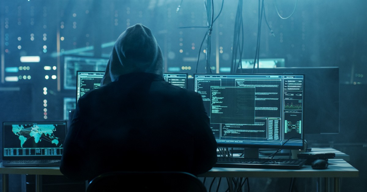 Indivíduo sentado em frente a múltiplas telas de computador em um ambiente escuro, com códigos e mapas globais exibidos nas telas, representando atividades de hacking ou segurança cibernética (cibercriminosos, hacker, cibercrimes, hp crimes cibernéticos, setor financeiro, finanças, bancos, dados, man-in-the-middle)