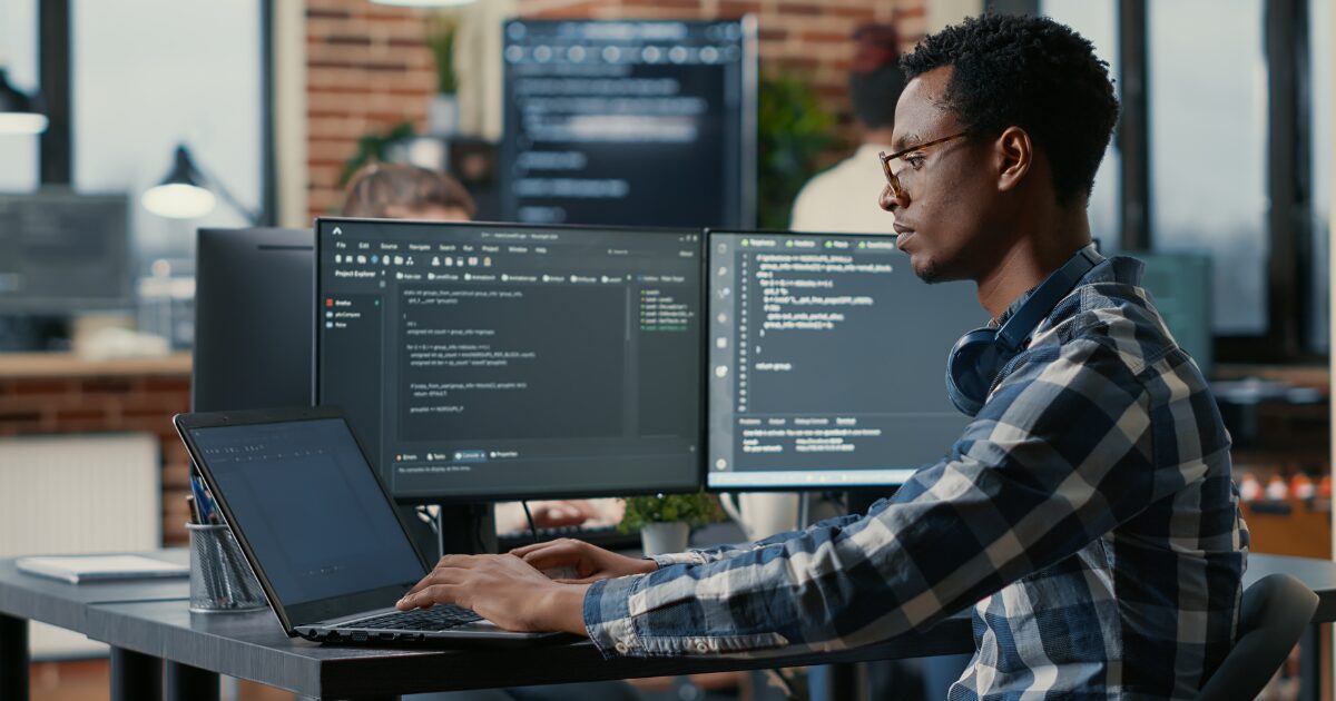 Homem negro usando óculos e camisa xadrez azul trabalha em um escritório moderno com três telas à sua frente, incluindo um notebook e dois monitores exibindo códigos de programação. Ele está concentrado no desenvolvimento de software, com fones de ouvido apoiados no pescoço. Ao fundo, há outros profissionais e um ambiente com tijolos aparentes e janelas amplas (desenvolvedores, OutSystems, desenvolvedores de software, Husky, cibersegurança, profissionais, software)