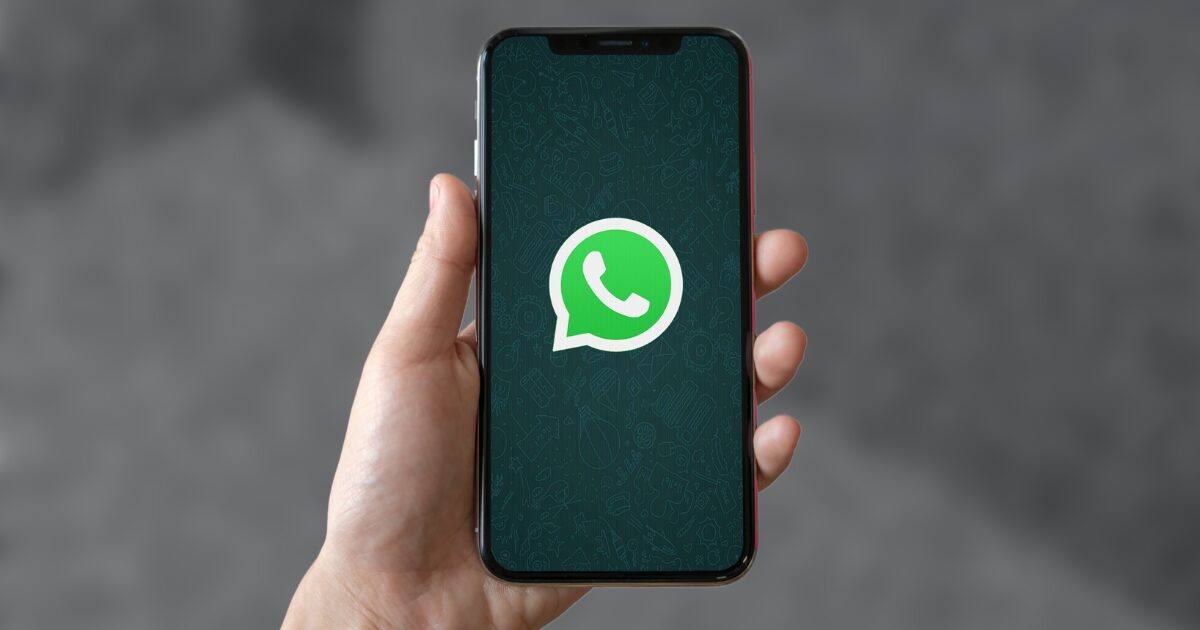 Imagem de uma mão segurando um smartphone que exibe a tela inicial do WhatsApp, com o ícone verde característico do aplicativo. O fundo desfocado em tons de cinza destaca o dispositivo e o logotipo do WhatsApp (whatsapp, mensagens, mensageria, varejo, rd station, Windows)