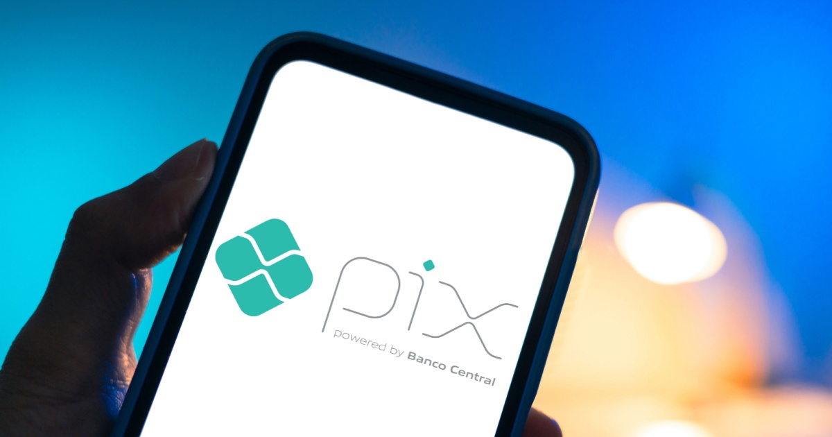 Pessoa segurando um smartphone que exibe na tela o logo do Pix, sistema de pagamento instantâneo brasileiro desenvolvido pelo Banco Central. O logotipo é composto por um símbolo geométrico verde ao lado da palavra "Pix" em letras estilizadas, seguido da frase "powered by Banco Central". O fundo da tela é branco e o ambiente ao redor tem iluminação com tons de azul e laranja desfocados (Sensedia)