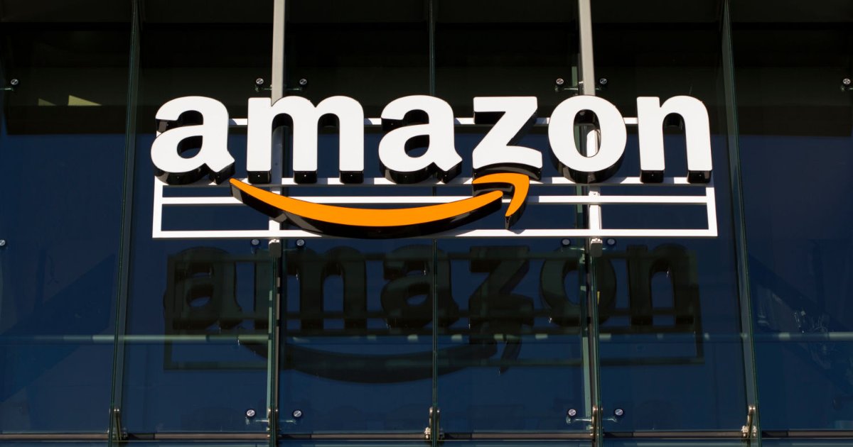 A imagem mostra o logotipo da Amazon em destaque, instalado na fachada de um prédio. O logotipo é composto pela palavra "amazon" em letras brancas, com a característica seta laranja que forma um sorriso abaixo da letra "a". A seta sugere a ideia de entrega rápida e satisfação. O fundo é formado por vidro reflexivo, o que permite que a imagem do logotipo seja refletida na superfície. A cena transmite um ambiente corporativo moderno e tecnológico (Amazon, dados, união europeia)