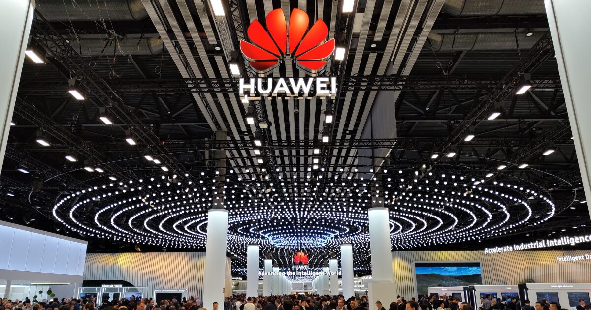 Estande da Huawei em um grande evento ou feira de tecnologia, com teto decorado por centenas de luzes em espiral e o logotipo da marca em destaque no centro superior da imagem. O local está lotado de pessoas, indicando grande interesse e movimentação no espaço da empresa. No fundo, é possível ver painéis e telas com informações tecnológicas, incluindo a frase "Accelerate industrial intelligence" (china, eua)