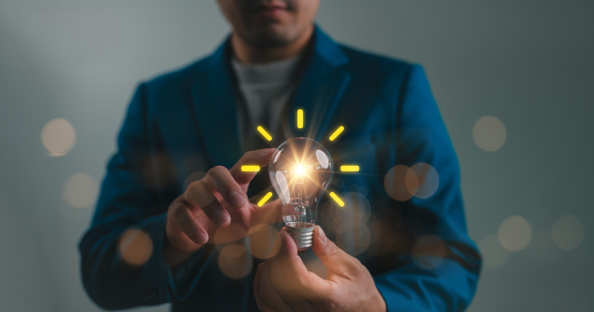Homem segurando uma lâmpada acesa com raios de luz emanando, simbolizando inovação e criatividade. Ele veste um blazer azul em um fundo neutro, destacando o conceito de ideias brilhantes e soluções inovadoras (inovação, empresa, startup, empresas, ideia, negócios, Andrade Gutierrez, tendências)