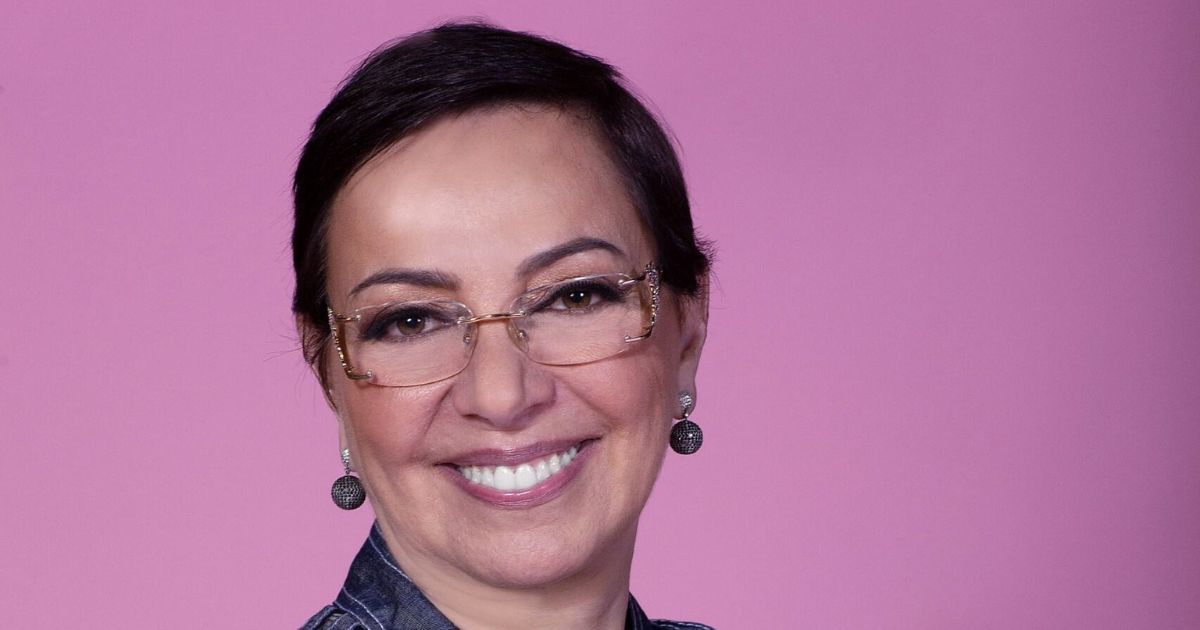 Tânia Cosentino, presidente da Microsoft Brasil, sorrindo para a câmera em um fundo rosa vibrante. Ela está usando óculos elegantes e brincos redondos, transmitindo uma imagem de simpatia e profissionalismo