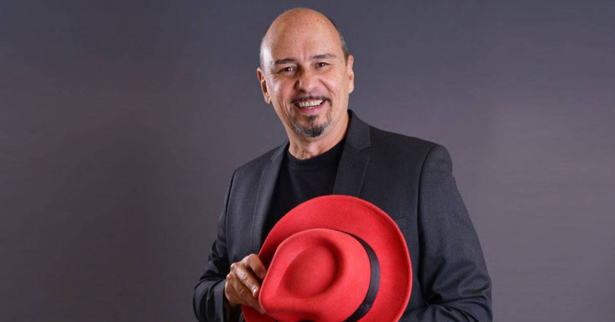 Na imagem, Gilson Magalhães, da Red Hat América Latina, tem uma expressão amigável, vestindo um blazer preto sobre uma camiseta da mesma cor. Ele segura um chapéu vermelho em uma das mãos, com um leve sorriso no rosto. O fundo é neutro, em um tom de cinza escuro, o que destaca ainda mais as cores da vestimenta e do chapéu. O chapéu vermelho é o ponto focal da imagem, adicionando um toque de cor vibrante que contrasta com o traje escuro. A pose e a expressão transmitem um ar confiante e acessível.