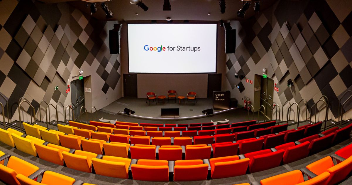 Auditório do Google for Startups, com assentos coloridos em tons de amarelo, laranja e vermelho, organizados em formato de arquibancada. Ao fundo, um grande telão exibe o logotipo 'Google for Startups', com cadeiras no palco para palestras. As paredes possuem design acústico geométrico (google for startups, google cloud)