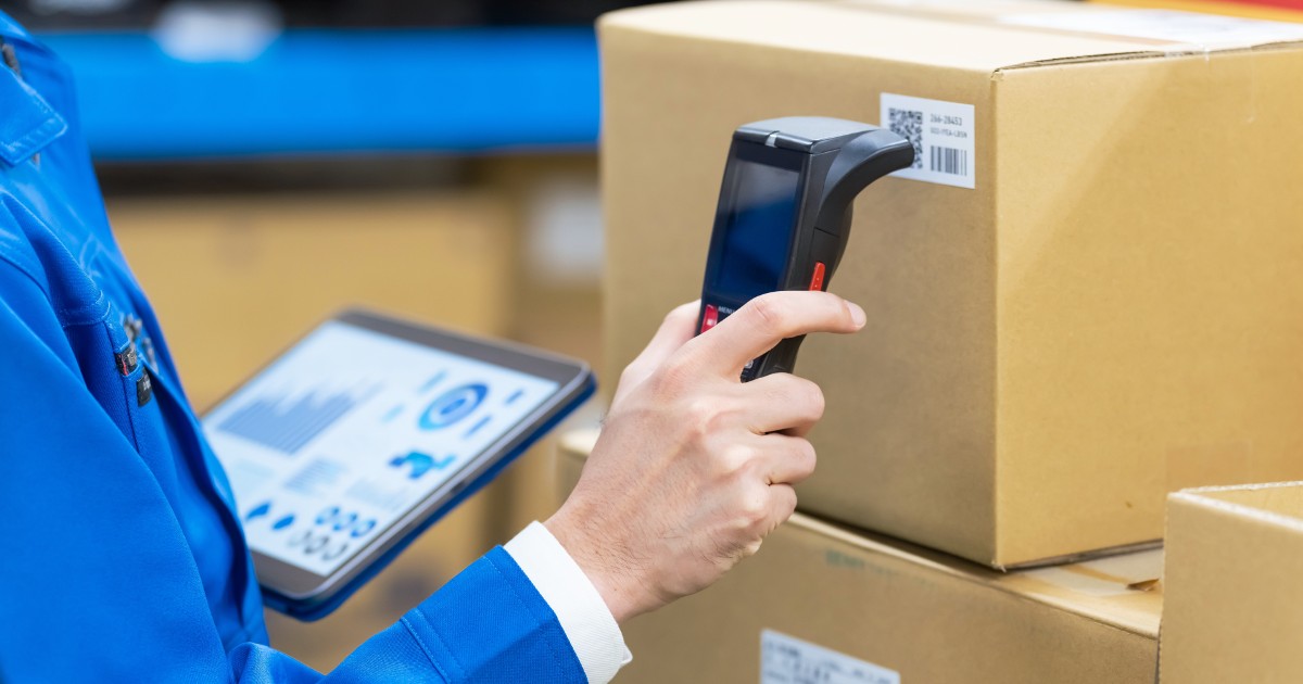 Trabalhador em um ambiente de logística utilizando um scanner de código de barras em uma caixa de papelão, enquanto segura um tablet com gráficos e dados exibidos na tela. Ele veste uniforme azul e está cercado por pacotes, sugerindo um processo de controle de estoque e rastreamento de produtos em um armazém (carbono)
