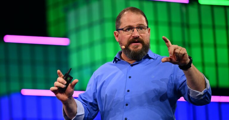 Cristiano Amon, presidente global da Qualcomm, falando no palco com gestos expressivos, usando uma camisa azul, com um fundo iluminado em tons vibrantes de verde e roxo