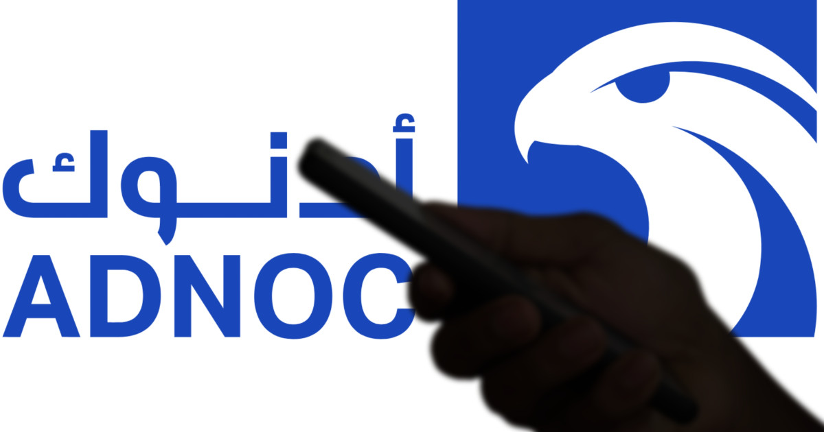 Logotipo da ADNOC, empresa de energia, com o icônico símbolo da águia em azul e branco, ao lado do nome da empresa em inglês e árabe. Em primeiro plano, uma silhueta de mão segurando um smartphone, sugerindo o uso de tecnologia ou comunicação digital relacionada à marca