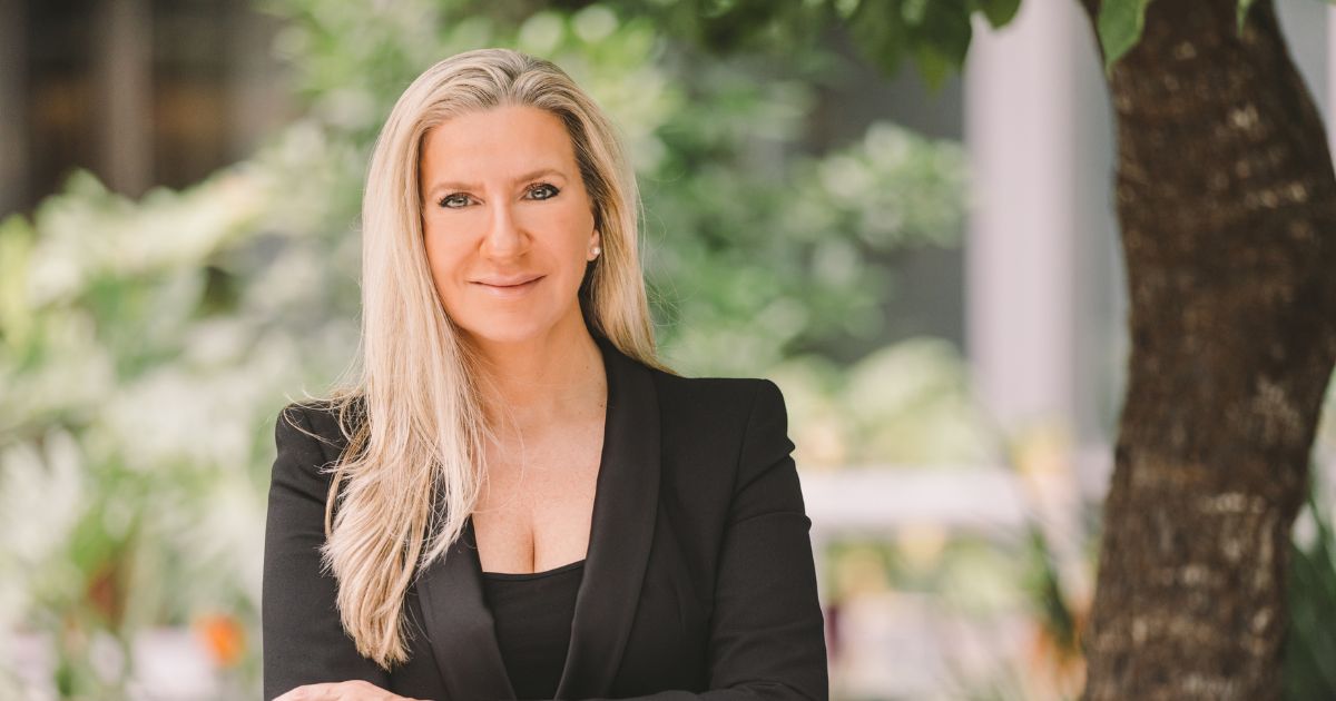 A imagem mostra Adriana Kersting, diretora de Recursos Humanos da SAP Labs, em um ambiente ao ar livre. Ela está usando um blazer preto, transmitindo uma postura profissional. O fundo desfocado destaca a vegetação, sugerindo um local com bastante natureza. Adriana está com os braços cruzados, exibindo confiança e simpatia, com um sorriso leve. A luz natural ilumina a cena, trazendo suavidade à composição.