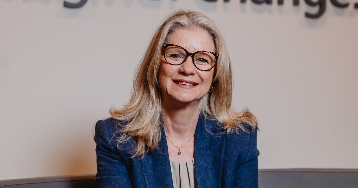 Andrea Klevenhusen, CEO da Ricoh Brasil, sorrindo enquanto está sentada. Ela usa um blazer azul e óculos de armação grande com cabelo loiro comprido e solto. O fundo é desfocado com um texto parcialmente visível