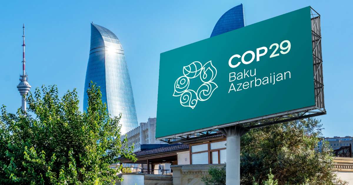 A imagem mostra um painel publicitário em Baku, Azerbaijão, com o logotipo do evento COP29. O painel tem um fundo verde e exibe o texto "COP29 Baku Azerbaijan", com um símbolo que representa o evento, possivelmente relacionado ao meio ambiente, em um estilo moderno. Ao fundo, são visíveis prédios altos e o céu claro. A imagem transmite a ideia de que Baku será a sede do COP29, um evento importante relacionado às mudanças climáticas