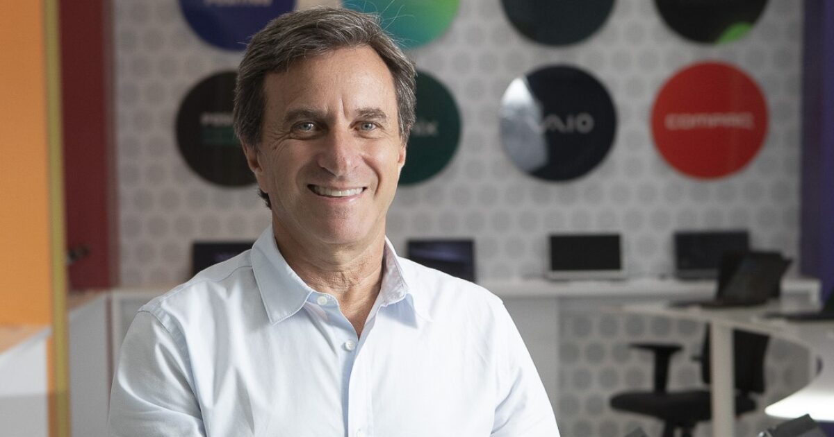 Hélio Bruck Rotenberg, presidente da Positivo Tecnologia, sorrindo para a câmera em um ambiente moderno com laptops ao fundo e placas coloridas decorativas, transmitindo uma imagem profissional e acolhedora. Ele veste uma camisa social branca