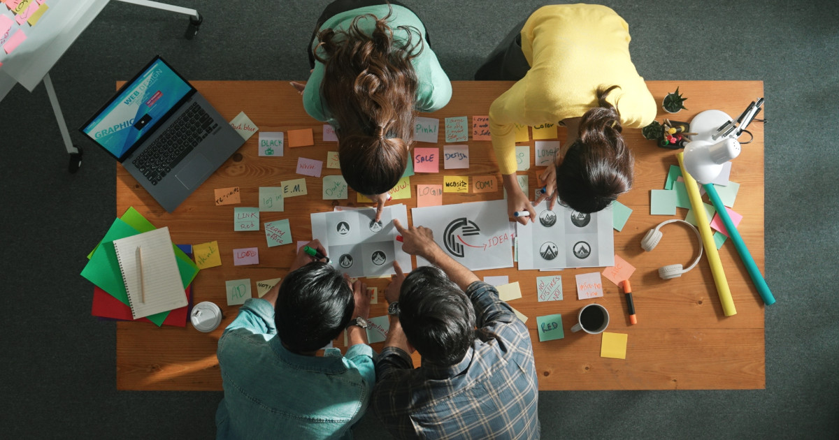 Imagem vista de cima de uma equipe colaborando em um projeto de design em uma mesa de madeira. Há papéis com esboços de logotipos, post-its coloridos espalhados com anotações, um laptop aberto, cadernos, canetas e outros materiais de escritório. A cena transmite criatividade, brainstorming e trabalho em equipe em um ambiente dinâmico