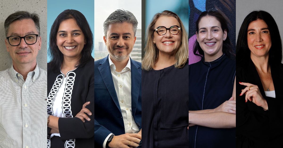 Retrato de seis executivos em um fundo neutro, mostrando diversidade de gênero e etnia. Eles estão posicionados lado a lado, cada um com uma expressão profissional e confiante. O primeiro executivo está usando óculos e camisa social clara, seguido por uma mulher de cabelos escuros e blazer preto com detalhes brancos. O terceiro é um homem de terno azul e camisa branca. Ao lado dele, uma mulher loira de óculos e blusa preta. A quinta pessoa é uma mulher sorridente com um fundo colorido e, finalmente, uma mulher de cabelos escuros e blazer preto fecha a imagem