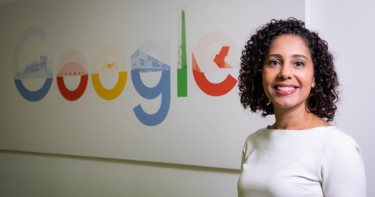 A imagem mostra Fernanda Doria, diretora de Negócios para Pequenas e Médias Empresas (PMEs) do Google. Ela está sorrindo e posando em frente a uma parede com o logotipo colorido do Google. Fernanda está usando uma blusa branca e brincos grandes, com seus cabelos cacheados soltos. O logotipo do Google na parede exibe cores vibrantes e elementos gráficos ilustrativos dentro das letras. O ambiente é de um escritório, transmitindo uma imagem profissional e inovadora