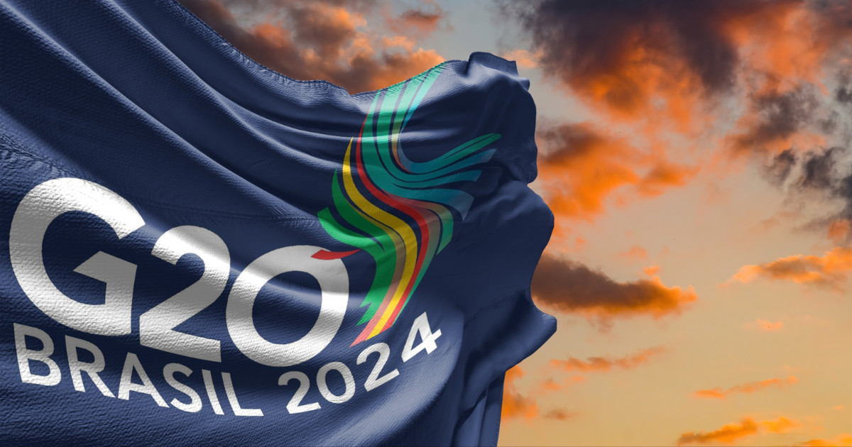 Imagem de uma bandeira azul escura com o logotipo do G20 Brasil 2024. O logotipo inclui a inscrição 'G20 Brasil 2024' em branco e um design colorido em forma de curva no centro, representando diversas cores vibrantes. Ao fundo, um céu com nuvens alaranjadas ao pôr do sol, criando um visual impactante e dinâmico (conservação ambiental)