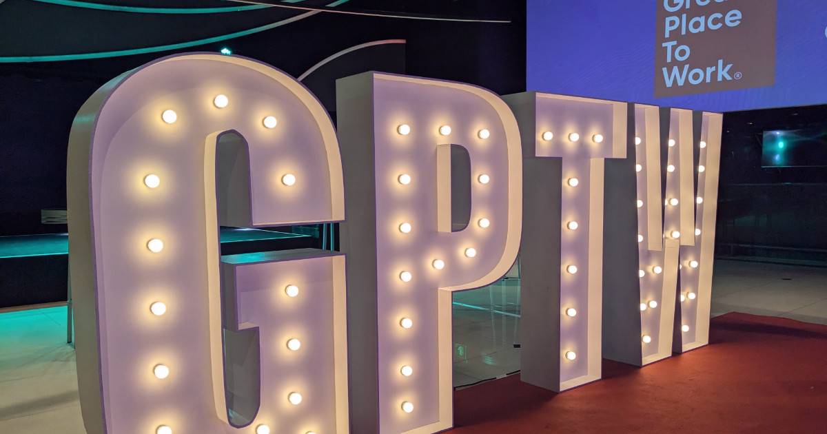 Imagem de grandes letras iluminadas formando a sigla 'GPTW', que significa 'Great Place to Work'. O cenário apresenta uma decoração moderna com luzes e projeções, destacando um ambiente corporativo ou evento relacionado a certificação de melhores empresas para se trabalhar GPTW 2024