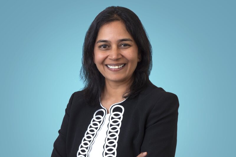 Retrato profissional de uma mulher de cabelos escuros, sorrindo e olhando diretamente para a câmera. Ela veste um blazer preto com detalhes brancos decorativos na parte frontal e posa com os braços cruzados. O fundo é azul claro e uniforme, destacando a figura, Lakshmi Hanspal DigiCert