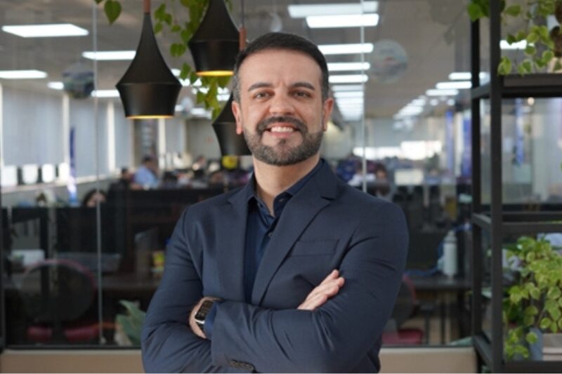 Retrato profissional de um homem de barba curta e escura, usando um terno azul escuro. Ele está sorrindo e posando com os braços cruzados em um escritório moderno, com luminárias suspensas e plantas ao fundo. A imagem mostra um ambiente de trabalho movimentado ao fundo, com pessoas em estações de trabalho, Marcio Marquese Simpress