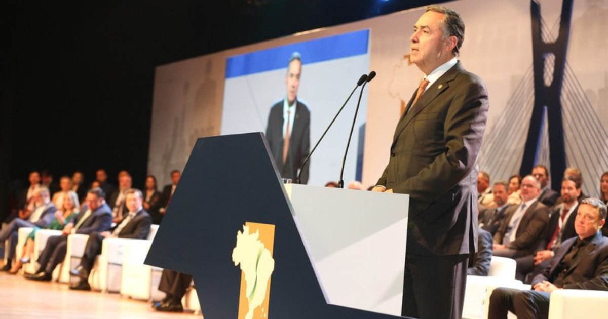 A imagem mostra um homem de terno, gravata e postura séria, discursando em um púlpito decorado com um mapa estilizado do Brasil. Ele está em um auditório, com diversas pessoas sentadas ao fundo, aparentando ser um público formal, possivelmente autoridades ou representantes de instituições. No palco, há um telão que exibe o orador, reforçando a ideia de um evento oficial ou conferência. Ao fundo, uma estrutura com design moderno, que pode ser associada à Ponte Estaiada, sugere que o evento ocorre em São Paulo ou remete à arquitetura da cidade. (STF)