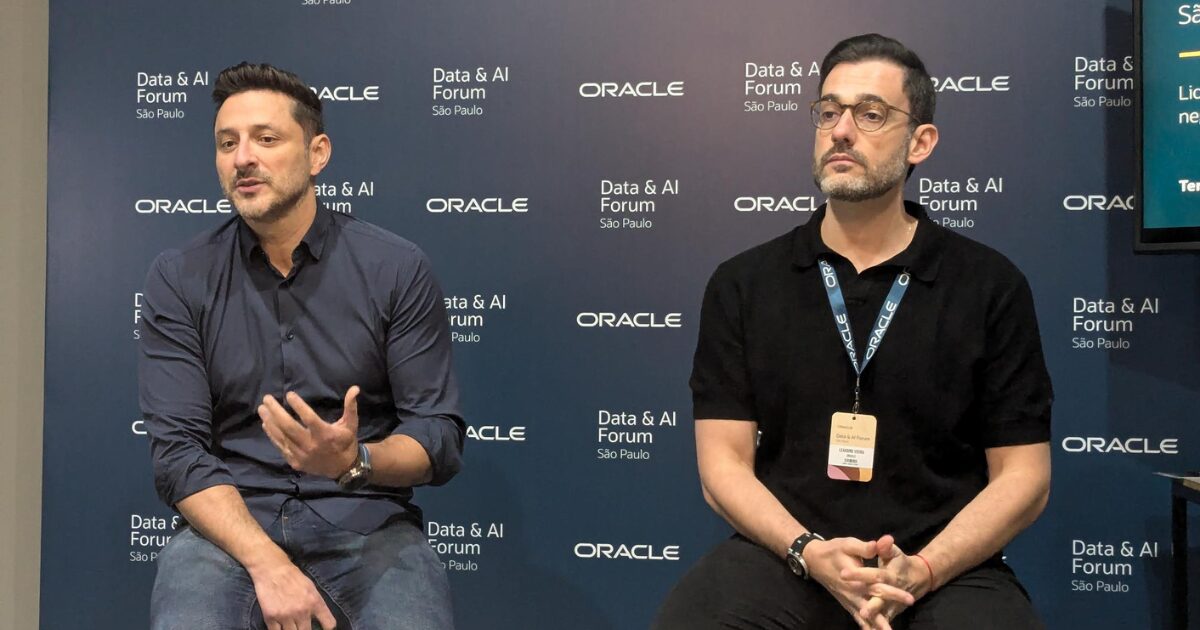Dois homens estão sentados em frente a um painel azul com o logotipo "Oracle" repetido, junto com o texto "Data & AI Forum São Paulo". O homem à esquerda está vestido com uma camisa social azul escura, gesticulando enquanto fala, com uma expressão engajada. O homem à direita usa uma camisa preta, óculos de grau e um crachá pendurado no pescoço, aparentando ouvir atentamente, com uma postura séria e mãos apoiadas no colo. A imagem parece capturada durante um evento ou painel de discussão sobre tecnologia e inteligência artificial