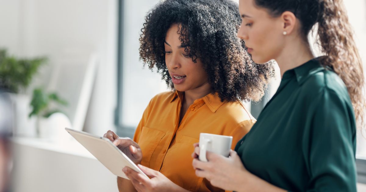 A imagem mostra duas mulheres em um ambiente de trabalho. A mulher à esquerda, que tem cabelo cacheado volumoso e usa uma camisa amarela, está segurando um tablet e explicando algo à outra mulher. A mulher à direita, que tem cabelo preso em um rabo de cavalo e veste uma camisa verde-escura, está segurando uma caneca branca enquanto observa atentamente a tela do tablet. O cenário ao fundo é iluminado, com tons claros, sugerindo um ambiente profissional ou de escritório.