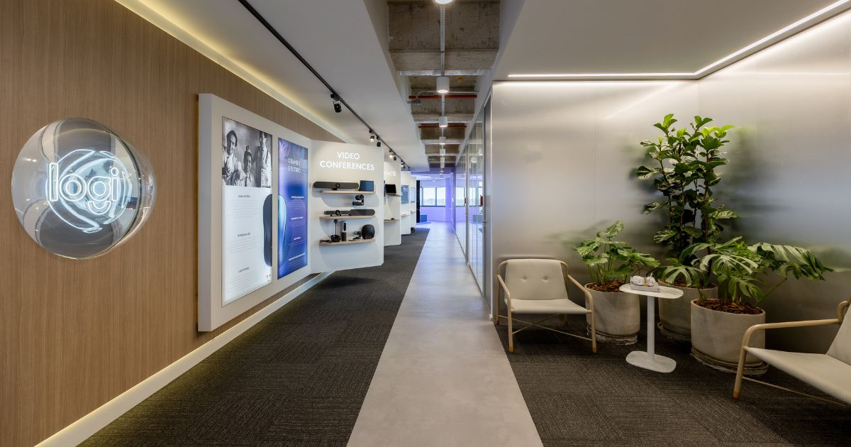 A entrada do showroom da Logitech em São Paulo transmite um ambiente moderno e sofisticado. À esquerda, o logo iluminado da "Logi" se destaca em uma parede de madeira clara, trazendo uma atmosfera acolhedora. Mais adiante, há uma seção de produtos voltados para videoconferência, exibidos em prateleiras suspensas e acompanhados por painéis com informações detalhadas, reforçando o compromisso da marca com tecnologia e inovação.
O corredor espaçoso segue em direção a áreas mais abertas, com iluminação indireta e design clean, combinando piso em concreto polido e carpete escuro. À direita, um espaço de espera confortável foi decorado com poltronas de design minimalista e vasos com plantas verdes, criando uma atmosfera de aconchego e frescor. Esse layout reflete a identidade da Logitech, unindo tecnologia e bem-estar em um ambiente funcional e esteticamente agradável.