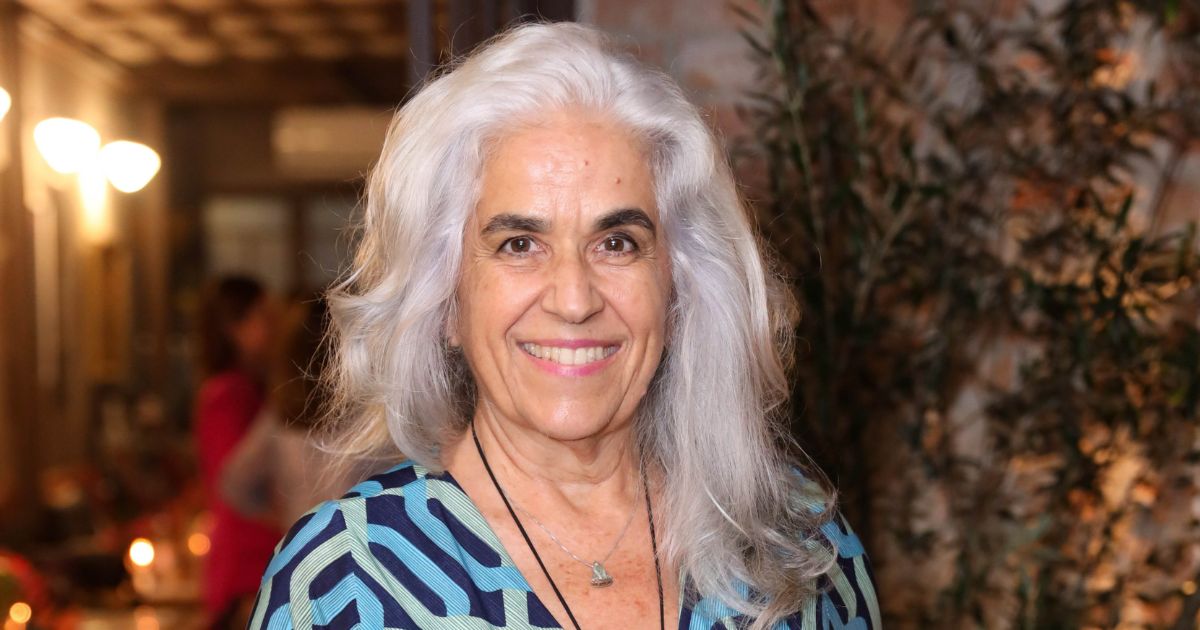 A foto mostra uma mulher de cabelos longos e grisalhos, com um sorriso gentil e caloroso. Ela veste uma blusa estampada em tons de azul e verde e usa um colar com um pingente delicado. Ao fundo, o ambiente é suavemente iluminado, com um toque de rusticidade, sugerido por plantas e luzes discretas. A expressão dela transmite simpatia e serenidade, destacando uma presença marcante e acolhedora.