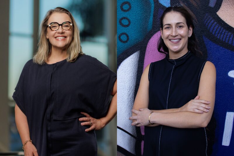 Retrato de duas mulheres lado a lado. À esquerda, uma mulher loira de óculos, usando uma blusa escura com mangas largas, sorri enquanto posa com uma mão na cintura. Ela está em um ambiente interno com janelas ao fundo. À direita, uma mulher de cabelos escuros vestindo um top preto sem mangas posa com os braços cruzados em frente a uma parede colorida com arte de grafite, também sorrindo, Vivian Broge, Marcela Zitune, IOS