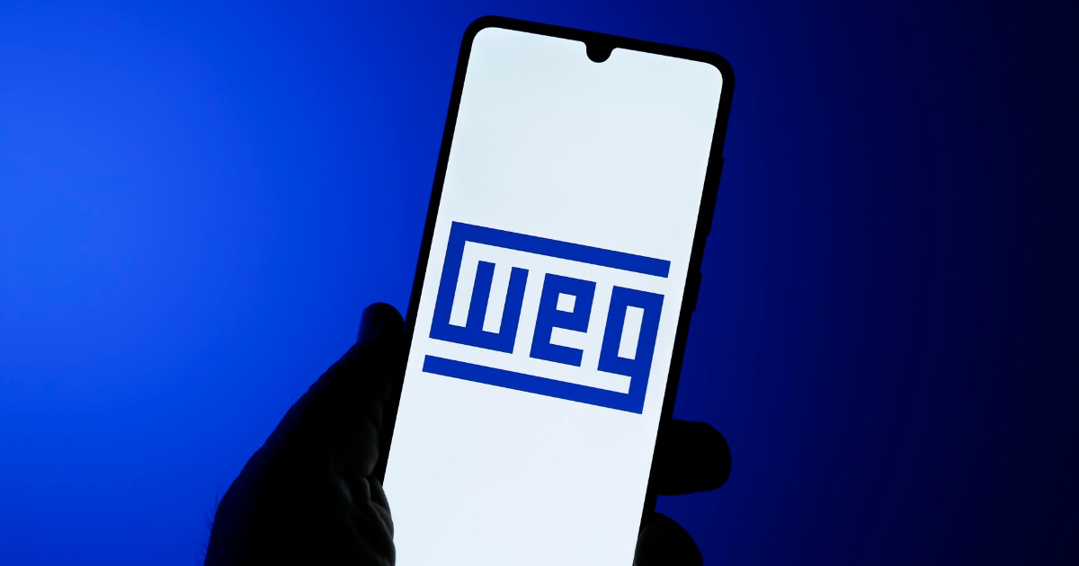 Logotipo da WEG exibido em um smartphone segurado por uma mão, com um fundo azul vibrante, simbolizando a tecnologia e inovação da empresa