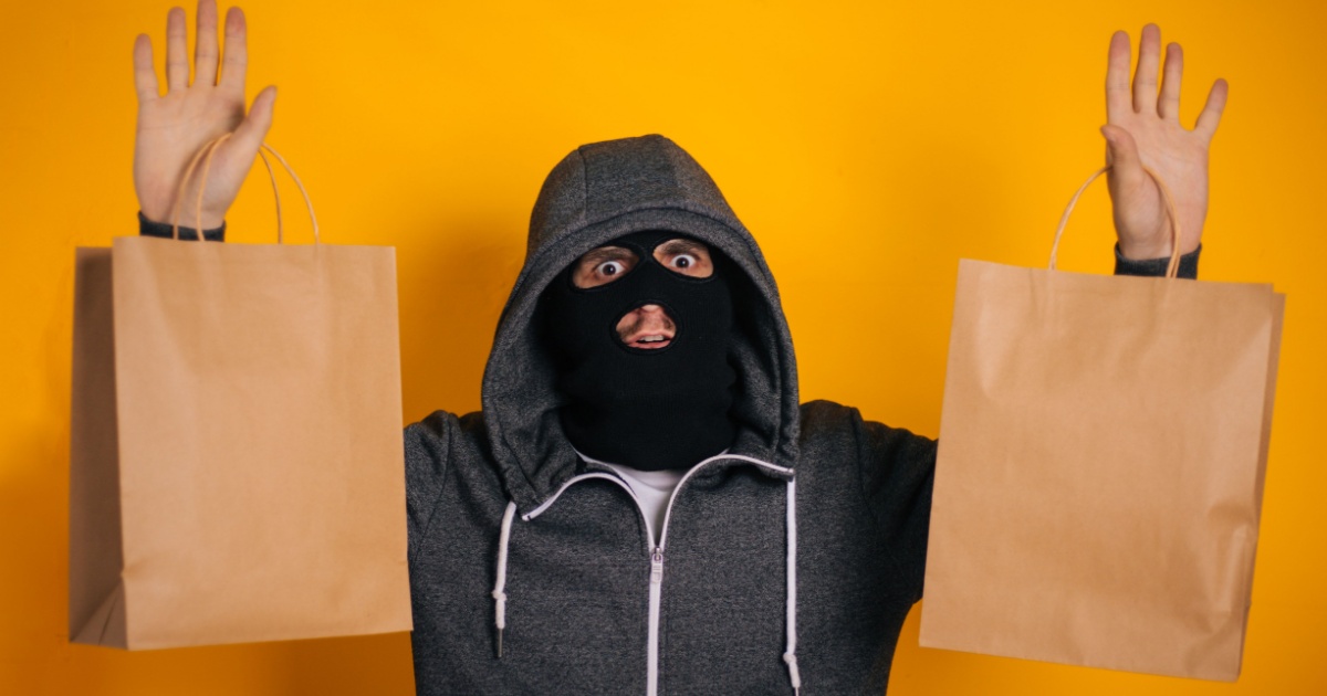Pessoa usando um moletom com capuz e uma máscara de ski preta, levantando as mãos e segurando sacolas de compras de papel kraft. A expressão facial parece surpresa ou assustada. O fundo é de cor amarela vibrante, destacando o contraste com o moletom cinza e a máscara preta (black friday, golpe, golpista, crime, criminoso, compras)