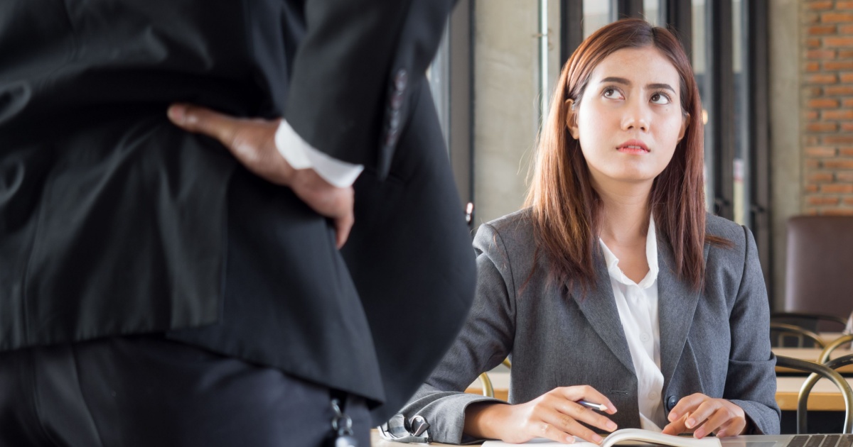Uma mulher de terno cinza, sentada e com expressão preocupada ou desconfiada, olhando para um homem em pé, parcialmente visível, que parece estar repreendendo ou confrontando-a. A cena sugere um ambiente tenso ou de confronto em um local de trabalho (chefe, cobrança, desconfiança, medo, assédio, trabalho, Slack)
