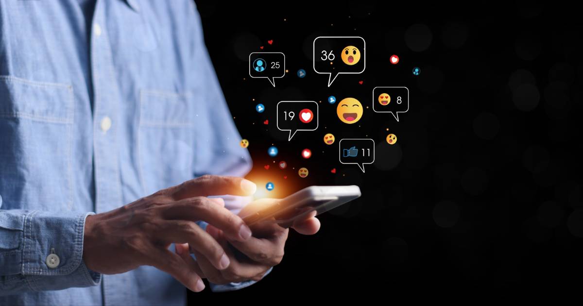 Pessoa utilizando um smartphone enquanto ícones de interações digitais, como curtidas, comentários e reações com emojis, flutuam ao redor da tela. O fundo escuro destaca a luminosidade dos ícones, representando o engajamento em redes sociais e o impacto da tecnologia na comunicação, abx, hiperpersonalização ( IA generativa), ABM