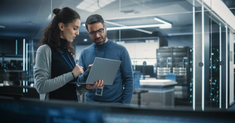 Dois profissionais de TI discutindo enquanto analisam informações em um laptop dentro de um data center moderno. O ambiente é iluminado por luzes azuis e cercado por servidores de alta tecnologia, com um cenário futurista e profissional, executivos, salários, empregos (Archimate)