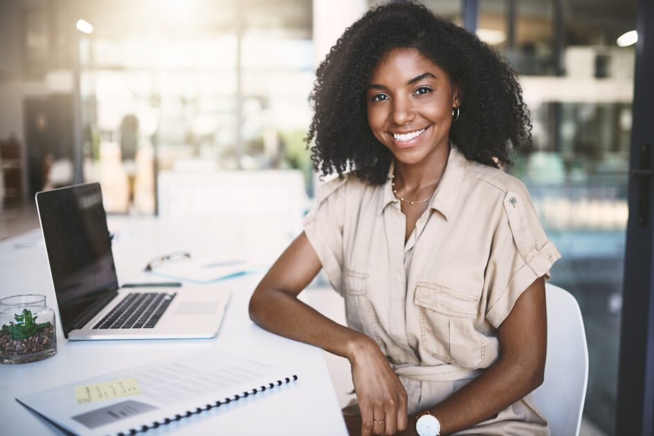 A imagem mostra uma mulher jovem com cabelo cacheado e sorriso amigável, sentada em um escritório moderno e iluminado. Ela veste um macacão bege casual e elegante, está sentada à mesa com um laptop aberto ao seu lado, junto a alguns documentos e uma pequena planta decorativa. O ambiente ao fundo está desfocado, mas parece ser um espaço de trabalho colaborativo, com grandes janelas e luz natural entrando. A expressão da mulher transmite confiança e simpatia.