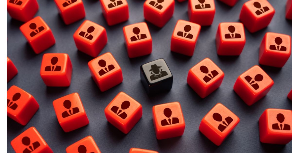 A imagem apresenta diversos cubos vermelhos com o ícone de uma figura de pessoa usando terno, representando executivos ou figuras corporativas. No centro da composição, destaca-se um cubo preto com um ícone de um agente secreto ou espião, simbolizando uma figura infiltrada ou anômala em meio ao grupo uniforme. A disposição em um fundo preto cria contraste, enfatizando o tema de diferenciação ou vigilância. Essa imagem pode remeter a conceitos como cibersegurança, espionagem ou ameaças internas em ambientes corporativos.