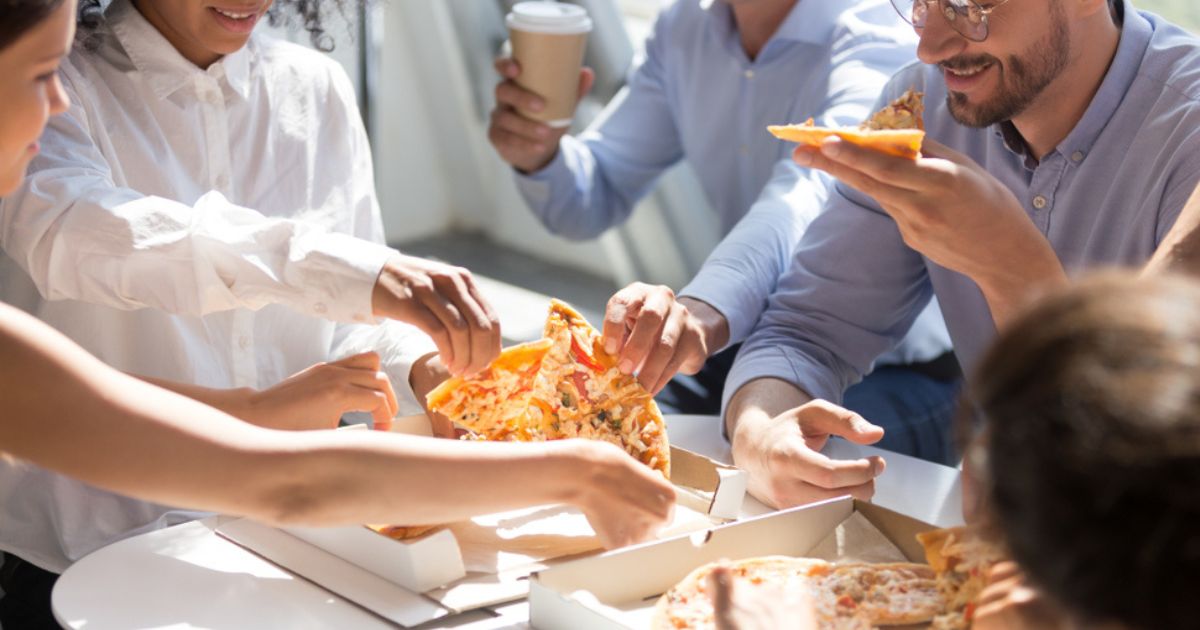 A imagem mostra um grupo de pessoas sentadas ao redor de uma mesa compartilhando pedaços de pizza em um ambiente iluminado, com luz natural entrando pelas janelas. Elas parecem estar em um momento descontraído e colaborativo, possivelmente em uma pausa no trabalho ou em uma reunião informal. Cada pessoa segura ou pega um pedaço de pizza, e o clima parece amigável e leve. A interação sugere um cenário de trabalho em equipe, com um tom descontraído, destacando a colaboração e a união.