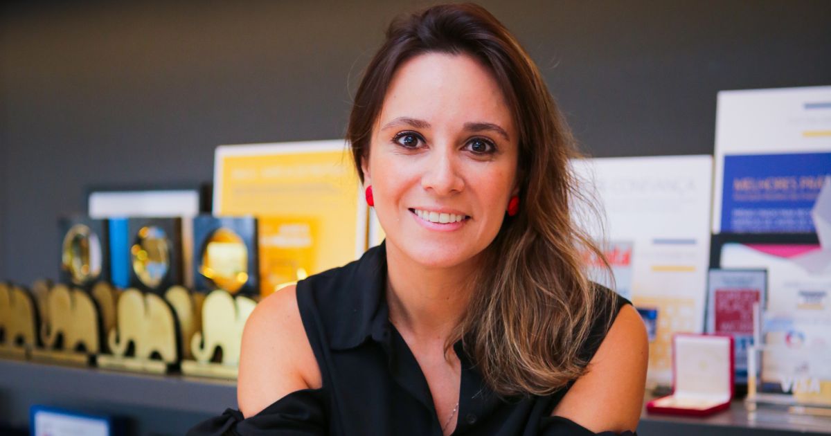 Uma mulher sorridente está sentada, olhando diretamente para a câmera. Ela tem cabelos castanhos claros, parcialmente soltos, e usa brincos vermelhos chamativos. Está vestida com uma blusa preta sem mangas. Ao fundo, há várias prateleiras decoradas com troféus dourados e placas de premiação, sugerindo um ambiente profissional e de conquistas.