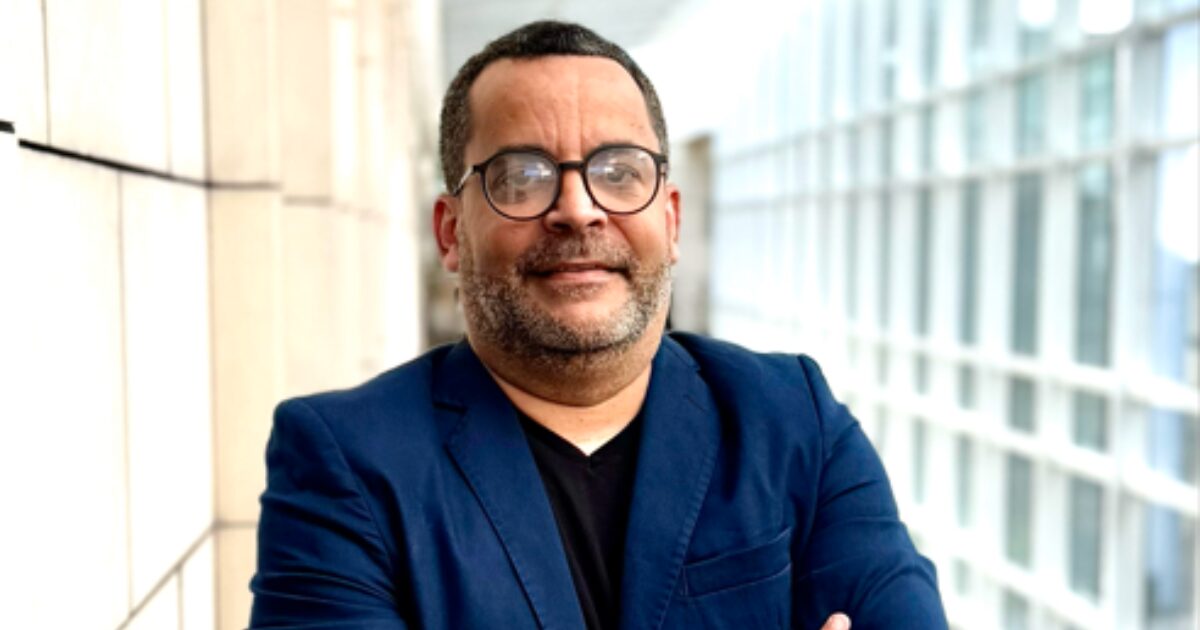 Um homem de pele clara, usando óculos de armação redonda, veste um blazer azul e está posicionado em frente a uma estrutura moderna de vidro e concreto. Ele sorri levemente, transmitindo confiança. A imagem ilustra um contexto sobre agentes de IA e inovação tecnológica.